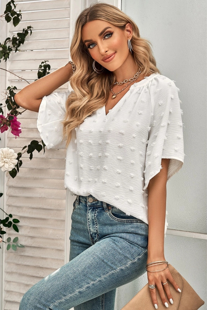 Woman wearing a white V-neck top with a textured pattern and batwing sleeves, paired with blue jeans. Also wearing jewelry, including a layered necklace, bracelets, and rings. 