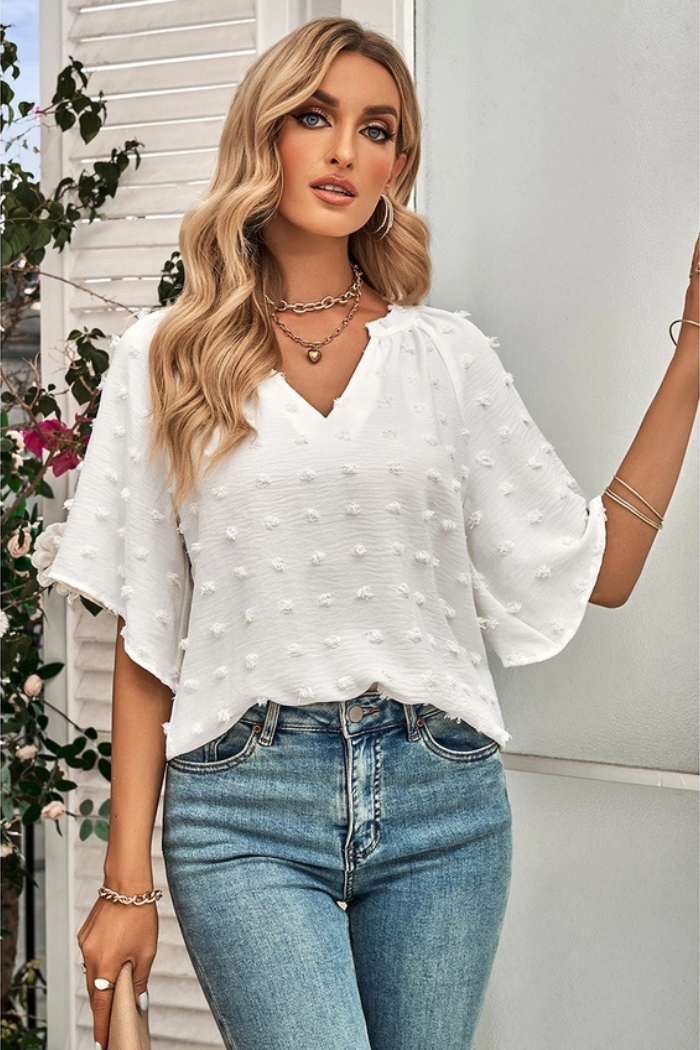 Woman wearing a white Swiss dot blouse with a notched neckline and flare sleeves. She is also wearing blue jeans. The blouse has a subtle texture due to the Swiss dots. She is accessorized with gold necklaces and bracelets. The background appears to be a light-colored wall with some greenery visible on the left side.