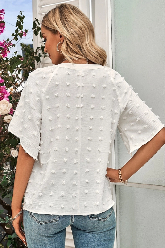Woman wearing a white shirt with a textured design and light-wash blue jeans. The shirt has short, flared sleeves and a V-neck. The woman is standing in front of a window with plants and flowers visible. The lighting suggests it might be daytime. The overall style of the clothing appears casual and fashionable.