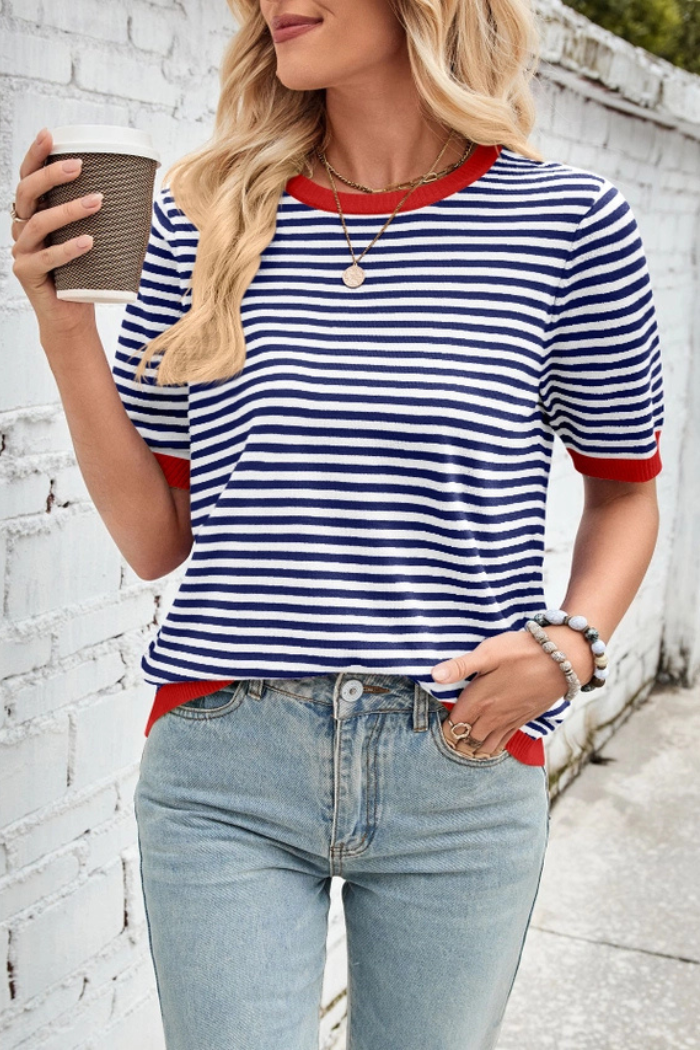 A woman wearing a navy and white striped short-sleeve knit top with a red trim on the collar and sleeves, paired with light blue jeans, holding a coffee cup.