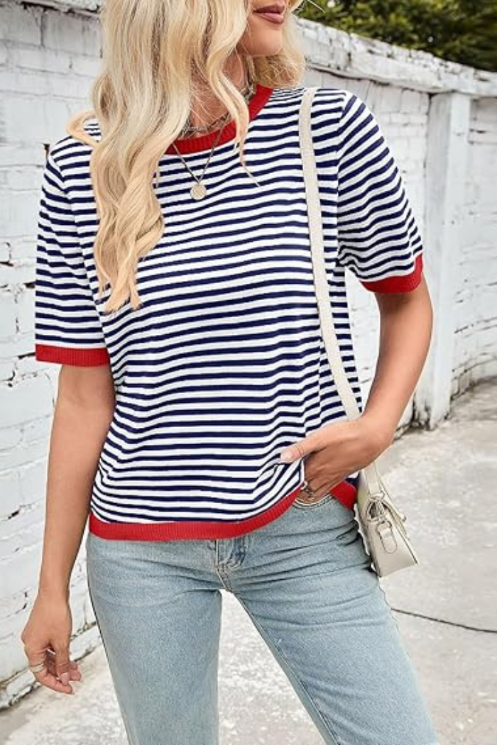 A woman with long blonde hair wears a navy and white striped short-sleeve t-shirt with red trim at the collar and sleeves, paired with light blue jeans and carrying a light-colored shoulder bag, standing outside against a light-colored brick wall.