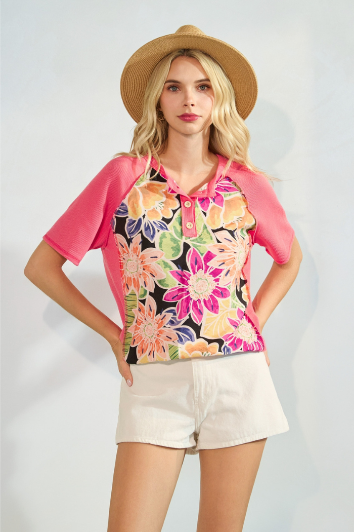 A blonde woman wearing a straw hat and a vibrant floral and pink short-sleeved top, paired with light-colored shorts, stands with her hands on her hips.