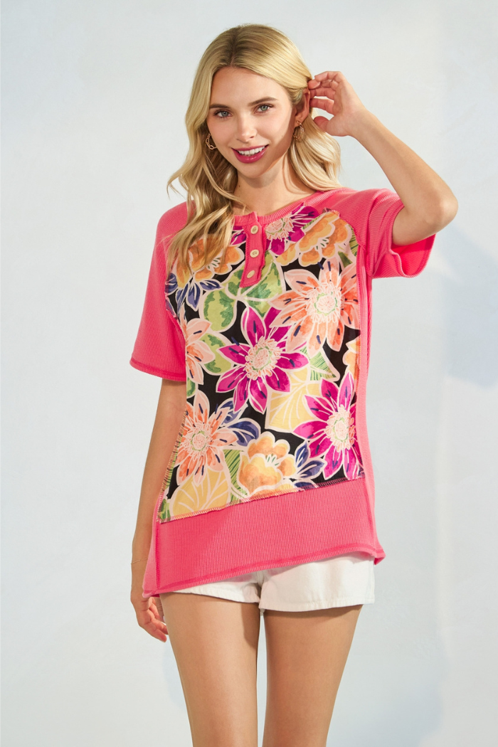A blonde woman smiles while modeling a coral short-sleeved top with a colorful floral panel on the front and white shorts. The top has a ribbed texture on the solid pink sections and a henley-style button placket at the neckline.