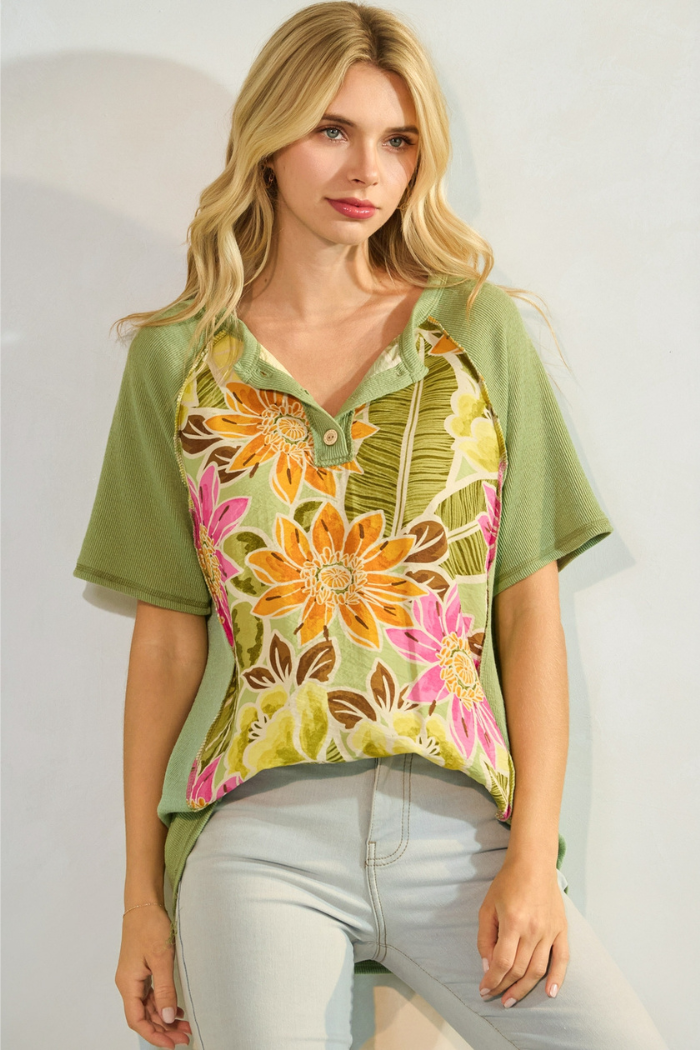 A young woman wearing a green and floral print short-sleeved top with a button-front Henley neckline and light-colored jeans.