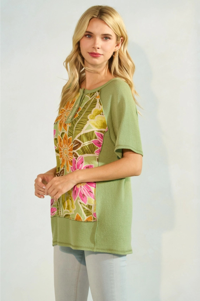 A woman with long blonde hair models a light green top with a floral print panel on the front. The floral panel features large pink, orange, and yellow flowers with green leaves on a lighter green background. The rest of the top, including the short raglan sleeves and the back, is a solid light green knit fabric. She is wearing light-colored pants.