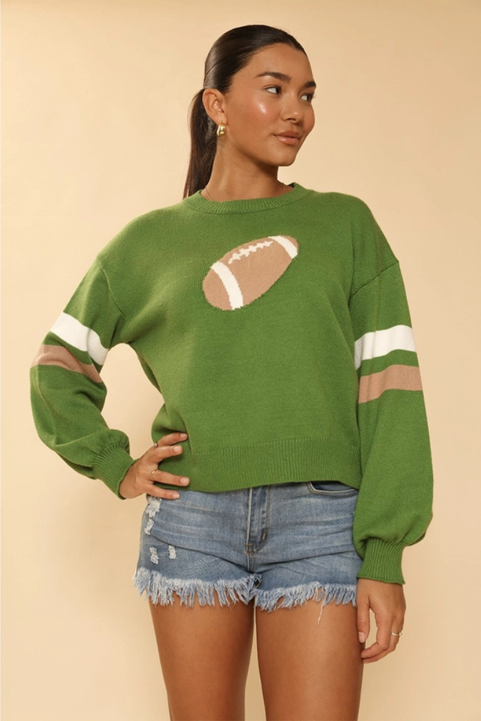 A woman wearing a green knit sweater with a football design on the front and white and brown stripes on the sleeves, paired with distressed denim shorts. 
