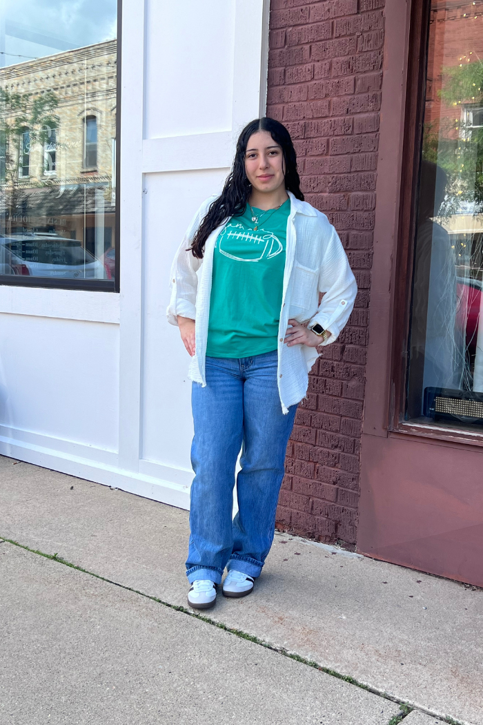 A young woman with long dark hair stands in front of a building with brick and white siding. She is wearing a green t-shirt with a football graphic, a white button-up shirt unbuttoned over it, blue jeans, and white sneakers. She has her hands on her hips and is looking directly at the camera.