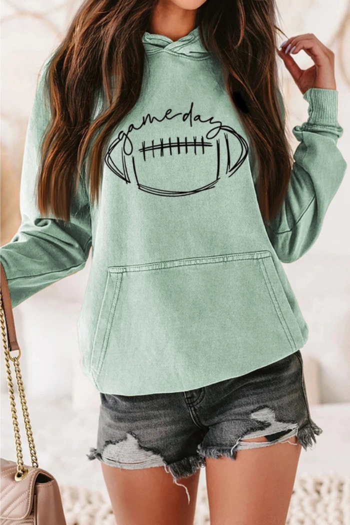 A light green long-sleeved hoodie with a large black graphic on the front featuring a football and the text "Game Day". The model is also wearing dark distressed denim shorts and carrying a light-colored chained shoulder bag. 