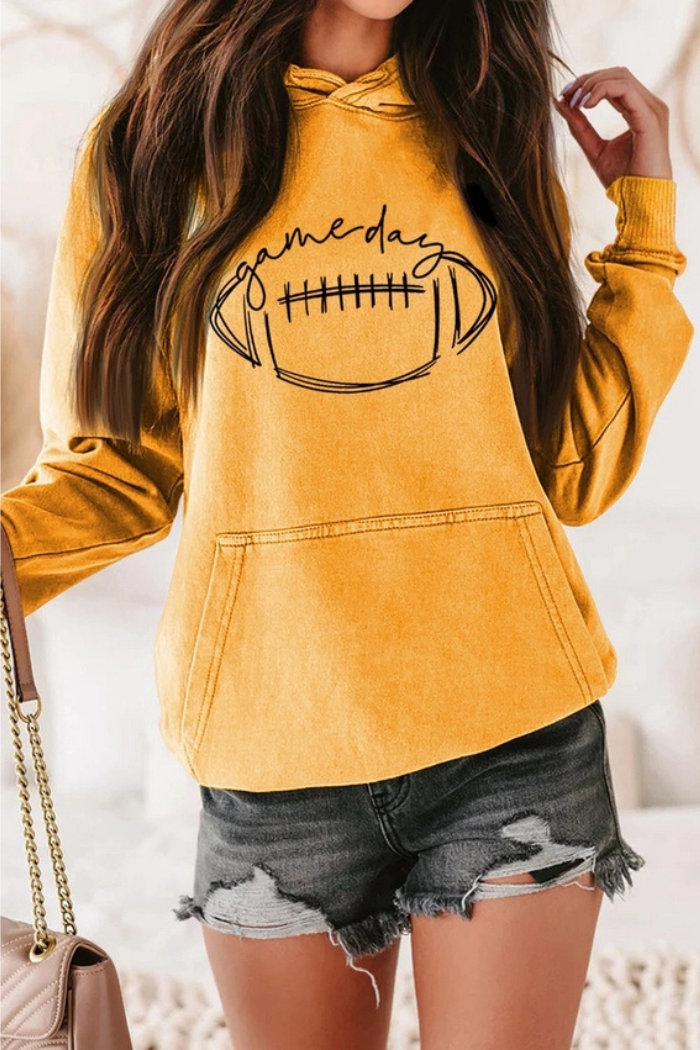 A Yellow long-sleeved hoodie with a large black graphic on the front featuring a football and the text "Game Day". The model is also wearing dark distressed denim shorts and carrying a light-colored chained shoulder bag. 