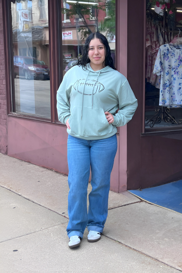 A young woman with dark, wavy hair stands on a sidewalk in front of a storefront, wearing a light green hooded sweatshirt with a football graphic and blue jeans, looking directly at the viewer.