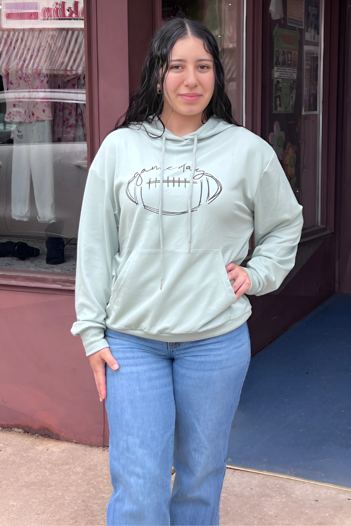 A young woman with dark, wavy hair stands on a sidewalk in front of a storefront, wearing a light green hooded sweatshirt with a football graphic and blue jeans, looking directly at the viewer.