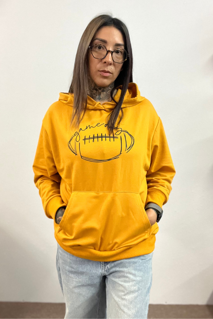 A woman with dark hair and glasses stands facing forward, wearing a mustard yellow hooded sweatshirt with a black outline drawing of a football and the word "gameday" written above it. The sweatshirt has a front pocket, and the woman's hands are in the pockets. She is also wearing light-colored jeans. The background is a plain white wall. 