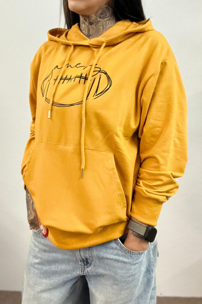 A person wearing a yellow hoodie with a black outline of a football and the word "gameday,." written above it, paired with light wash jeans. The person has tattoos visible on their neck and hands and is wearing a dark-colored watch on their left wrist.