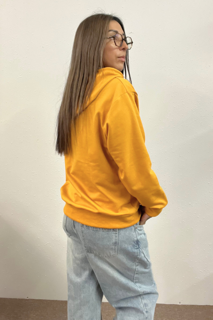 A woman wearing a solid yellow hoodie and light-wash, loose-fitting jeans is shown from a rear-side perspective, looking over her right shoulder. 