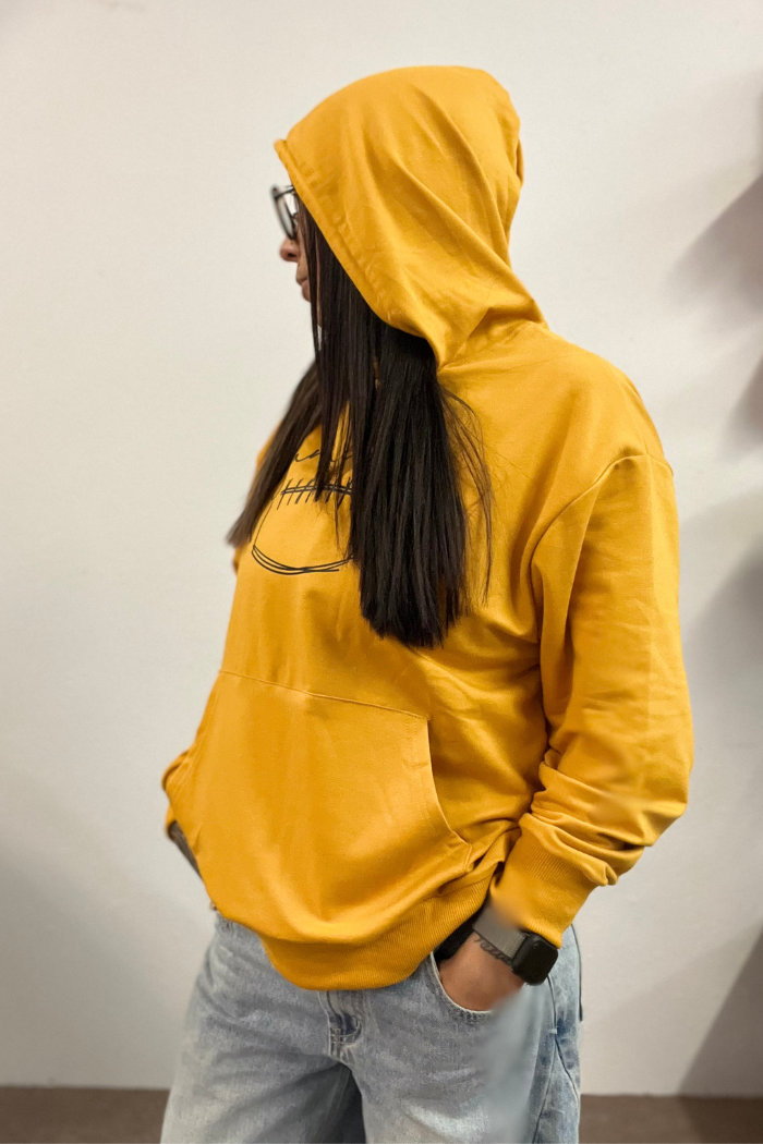 A person with long, dark hair and glasses wearing a yellow hoodie with a graphic print on the chest, standing with hands in the kangaroo pocket, viewed from the side.