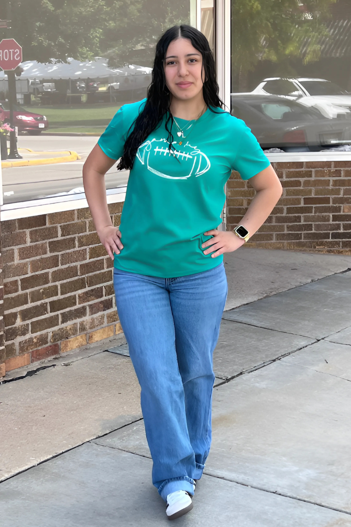 A young woman with long, dark hair stands outdoors, facing forward with her hands on her hips. She is wearing a teal t-shirt with a white outline of a football on the front, light blue wide-leg jeans, and white shoes. She is also wearing a necklace and a watch on her left wrist. The background features a brick wall, a window reflecting trees and a building, and a street with cars in the distance.