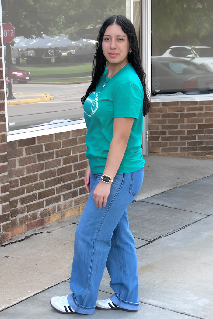 A young woman with long dark hair stands on a sidewalk, smiling slightly, wearing a teal t-shirt, blue jeans, and white sneakers with black stripes. A stop sign and reflections of cars and buildings are visible in the background.
