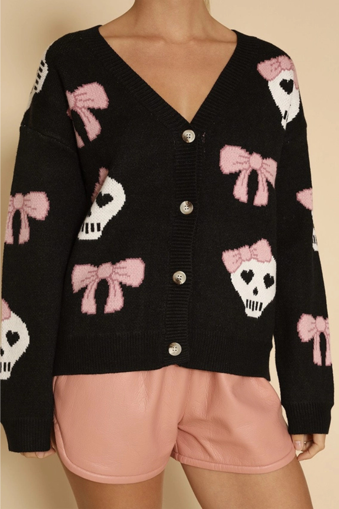 A woman wears a black v-neck cardigan with white skull and pink bow intarsia patterns, paired with light pink shorts.