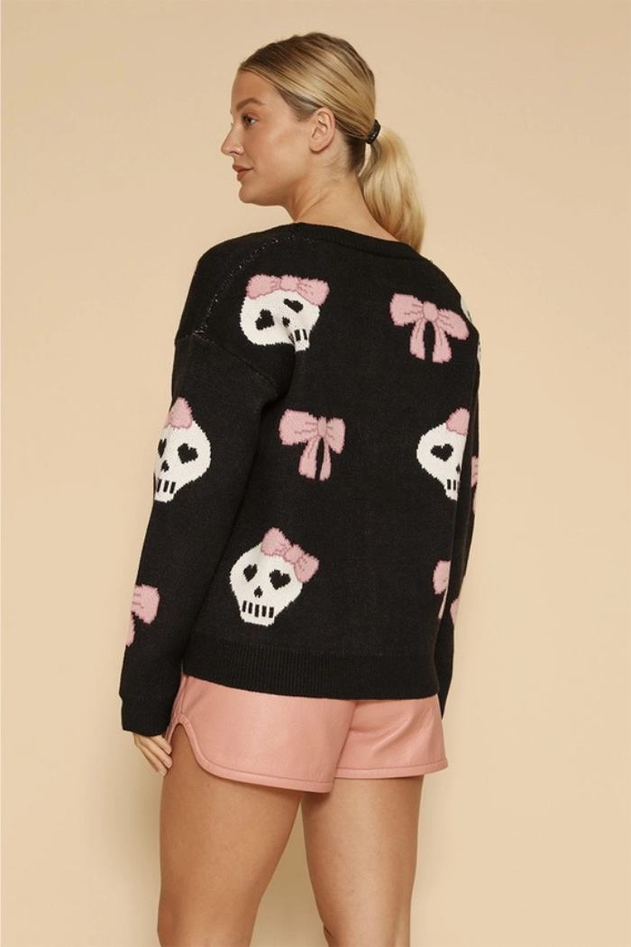 A woman stands with her back to the camera, wearing a black long-sleeved sweater adorned with white skulls featuring pink bows, and light pink shorts. Her blonde hair is tied back in a ponytail.