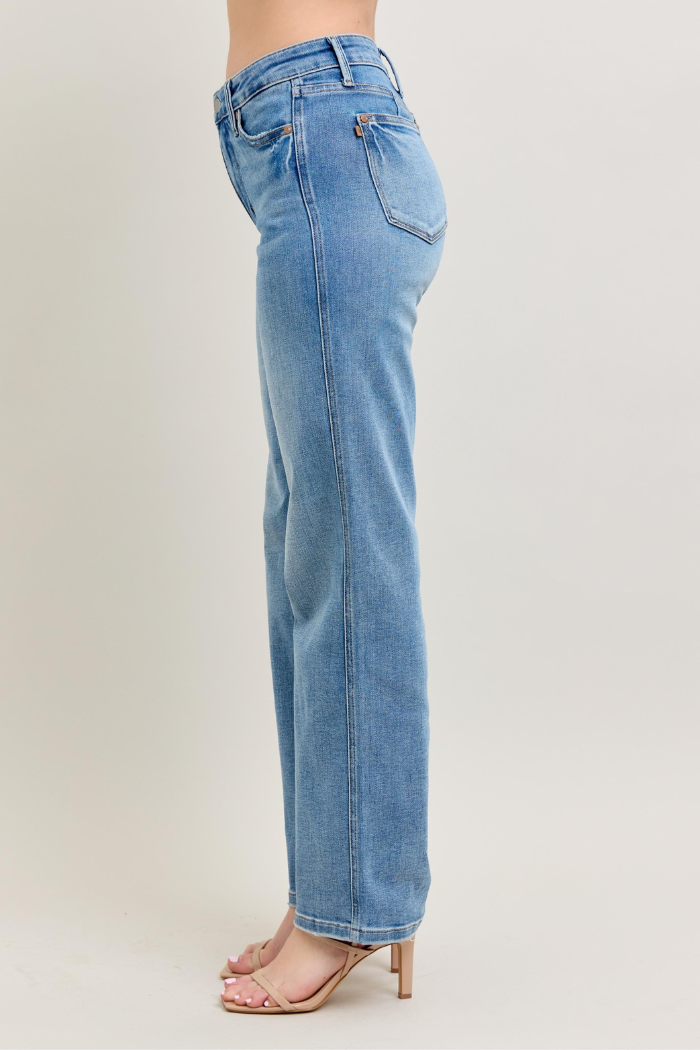 A side profile of a person wearing light wash, straight-leg Judy Blue jeans with visible tummy control details, paired with neutral-toned heeled sandals, on a light background.