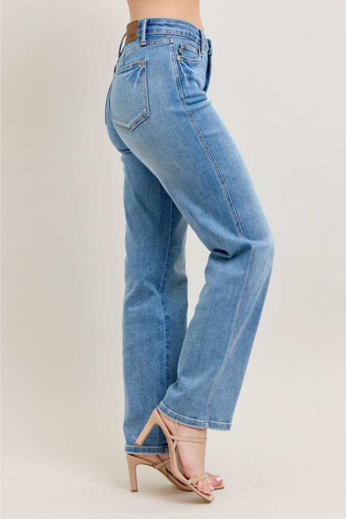 A side view of a person wearing light blue, vintage wash, straight-leg Judy Blue jeans with a visible back pocket and a mid-rise fit, paired with nude high-heeled sandals. 