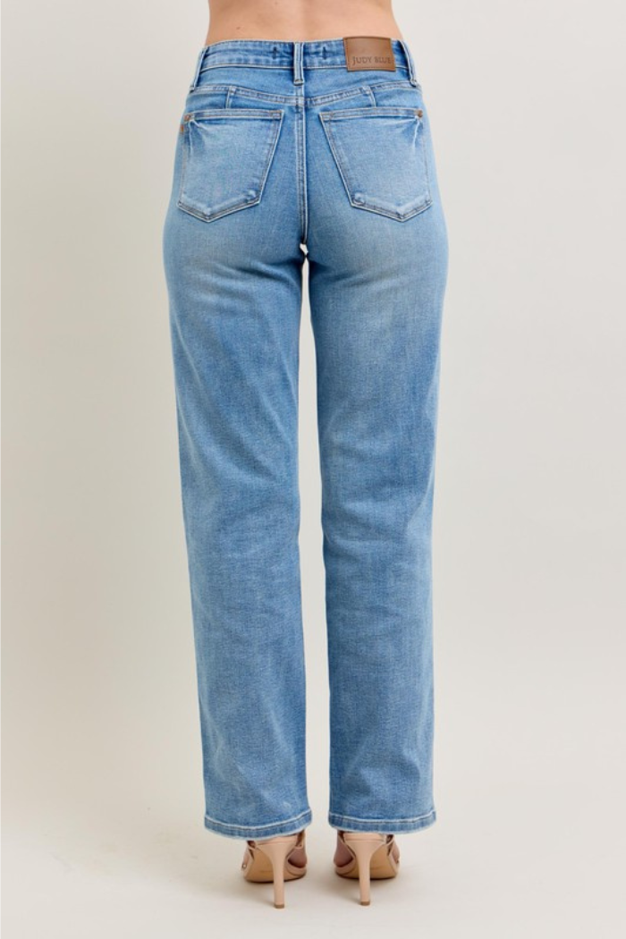 A pair of Judy Blue Mid Rise Tummy Control Vintage Wash Straight Jeans in a light blue wash, shown from the rear view, worn by a person standing on a white background.