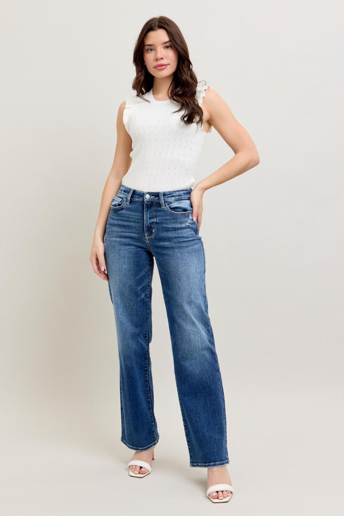 The image shows a woman wearing a white, sleeveless, ribbed knit top with ruffled details on the shoulders.
High-waisted, medium-wash blue jeans with a straight leg fit, likely from the brand Judy Blue. White open-toed sandals with a low block heel.
