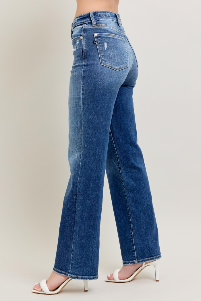 The image shows a pair of Judy Blue High Waist 90's Straight Leg Jeans in a dark blue wash, worn by a person from the waist down, facing slightly away from the camera.