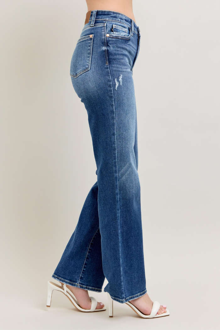 A side profile image of a person wearing dark wash, high-waist, 90's style straight leg jeans by Judy Blue, paired with white heeled sandals. The jeans show subtle distressing on the thigh.
