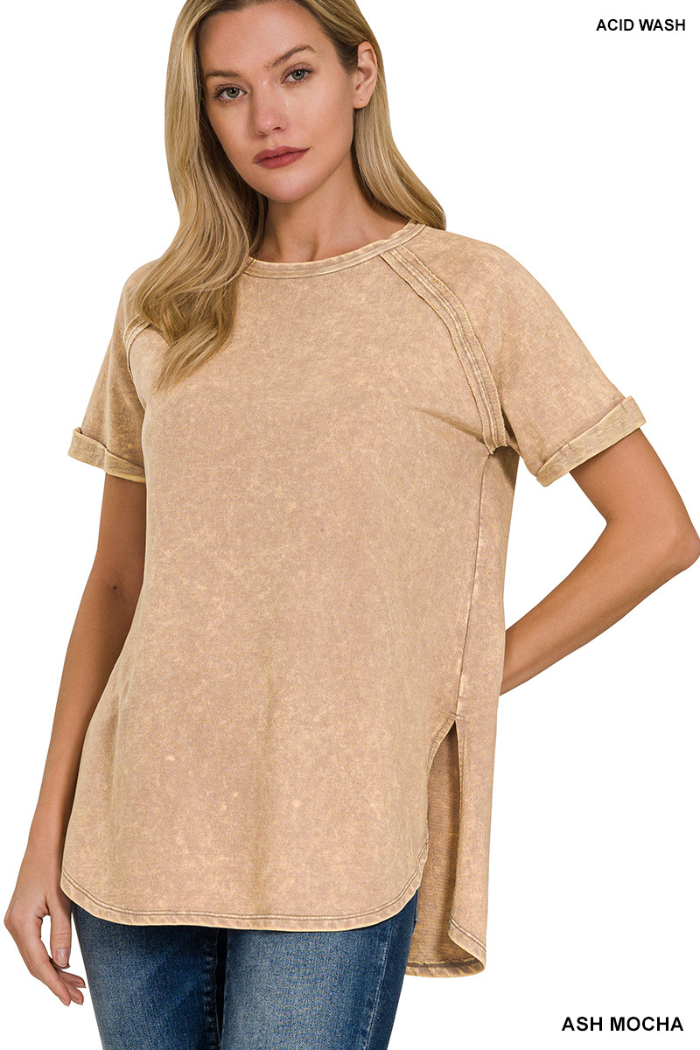 A woman wearing a relaxed-fit, short-sleeved, scoop-neck top in a light, vintage-washed or acid-washed tan/beige color. The top has visible stitching details on the shoulders and a rounded hem with side slits. The woman has long, blonde hair and is posing against a white background.