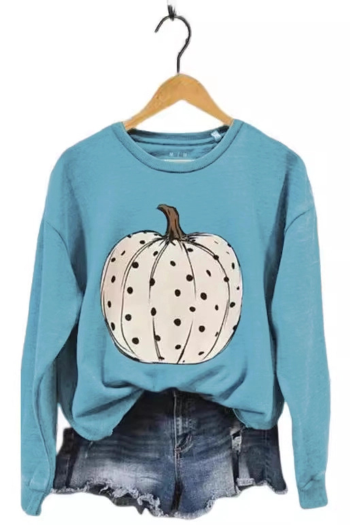 A light blue long-sleeved crewneck sweatshirt with a large white pumpkin on the front, covered in small black polka dots. The sweatshirt is displayed on a wooden hanger over a pair of denim shorts.