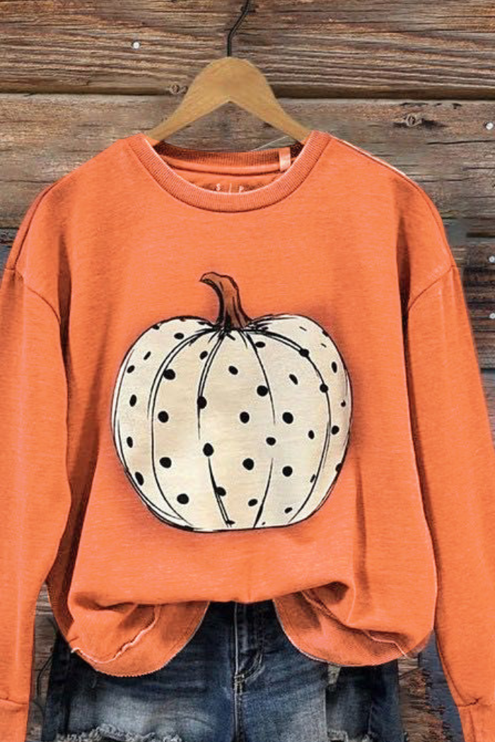 An orange long-sleeved crewneck sweatshirt with a large white pumpkin on the front, covered in small black polka dots. The sweatshirt is displayed on a wooden hanger over a pair of denim shorts.