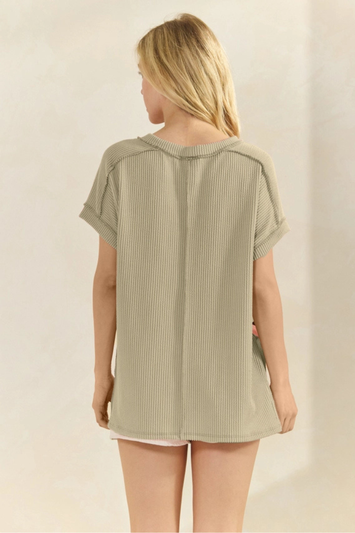 A woman, viewed from the back, wears a relaxed-fit, short-sleeved, ribbed knit top in a muted olive-green color. The top features a visible seam down the center back and appears to have rolled or cuffed sleeves. Her blonde hair is pulled up, and she stands against a light-colored background.