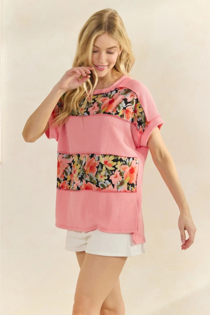 A blonde woman models a pink/coral, ribbed knit top with floral print panels across the chest and stomach, paired with white shorts. She is smiling and looking slightly down and to her right, with her left hand near her face.
