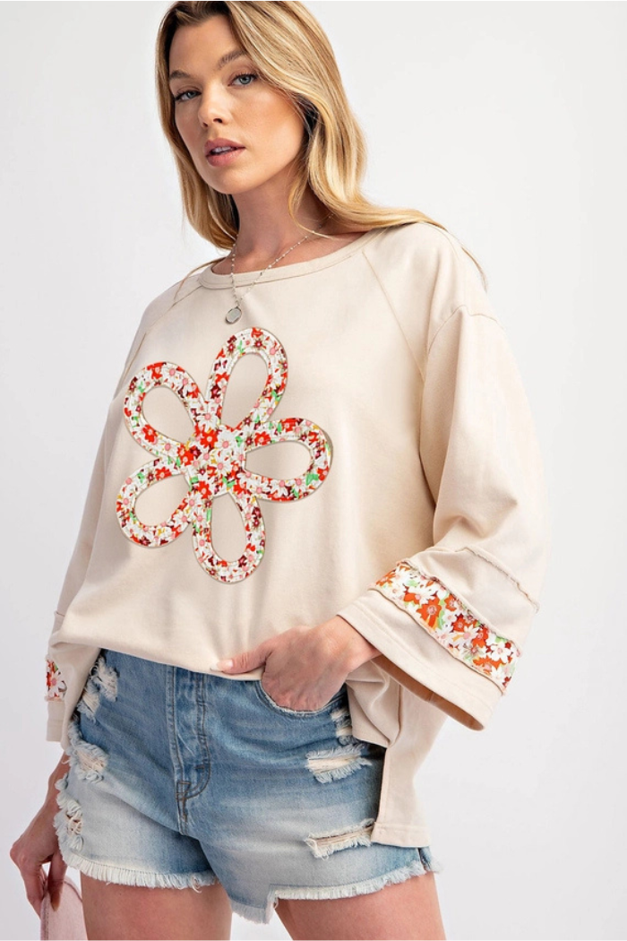 A woman wearing a beige long-sleeved top with a large, colorful floral appliqué on the front and matching floral cuffs, paired with distressed denim shorts.
