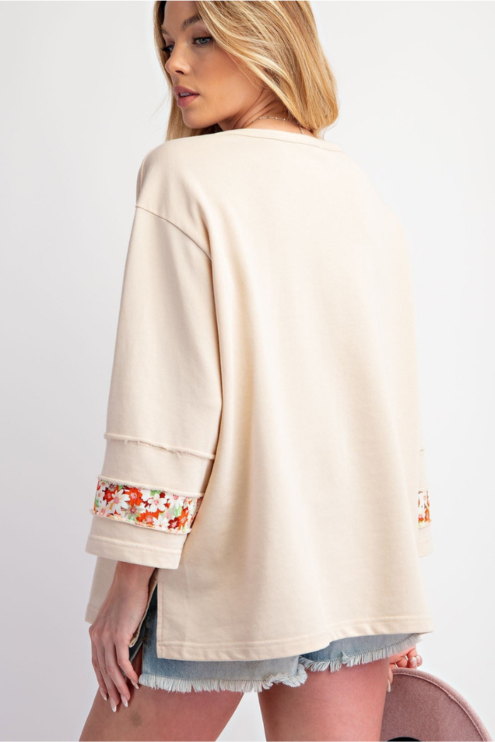 Back view of a woman wearing an oversized, cream-colored sweatshirt with floral pattern trim on the cuffs of the three-quarter length sleeves, paired with denim shorts.

