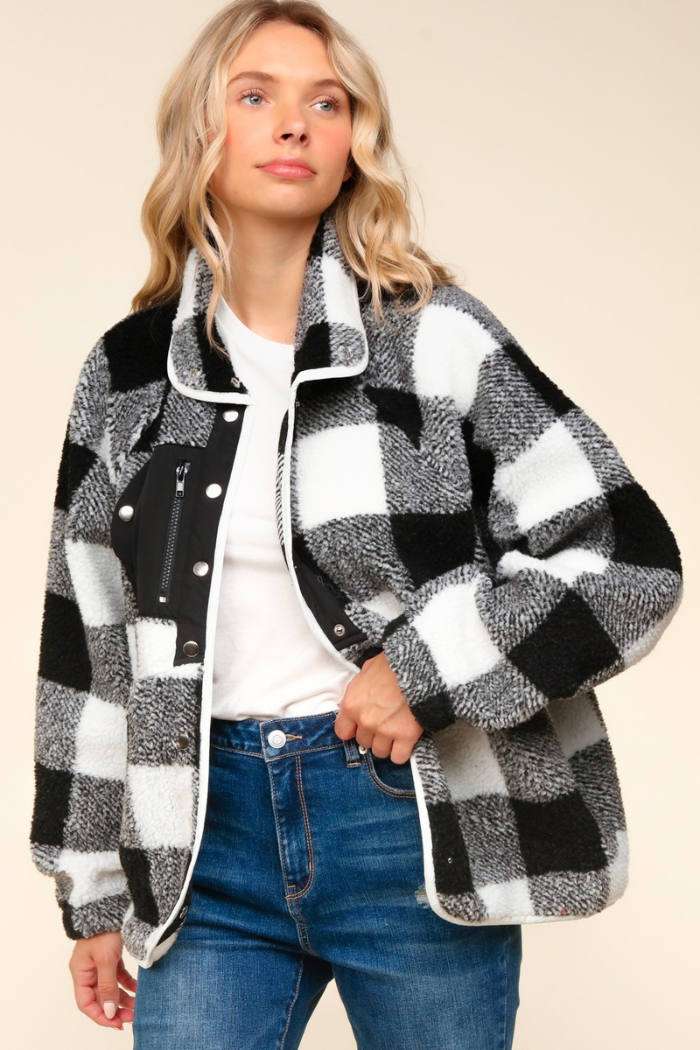 A woman with blonde hair wears a black and white buffalo plaid sherpa jacket over a white t-shirt and blue jeans. The jacket has a high collar, a zippered pocket on the left chest, and snap-button closures.