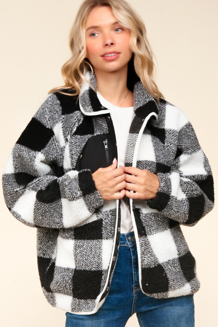 A blonde woman models a black and white buffalo plaid sherpa fleece jacket with a high collar, a black zippered chest pocket, and a full-zip front closure with white trim. The jacket features long sleeves and a relaxed fit, and the woman is wearing blue jeans. 
