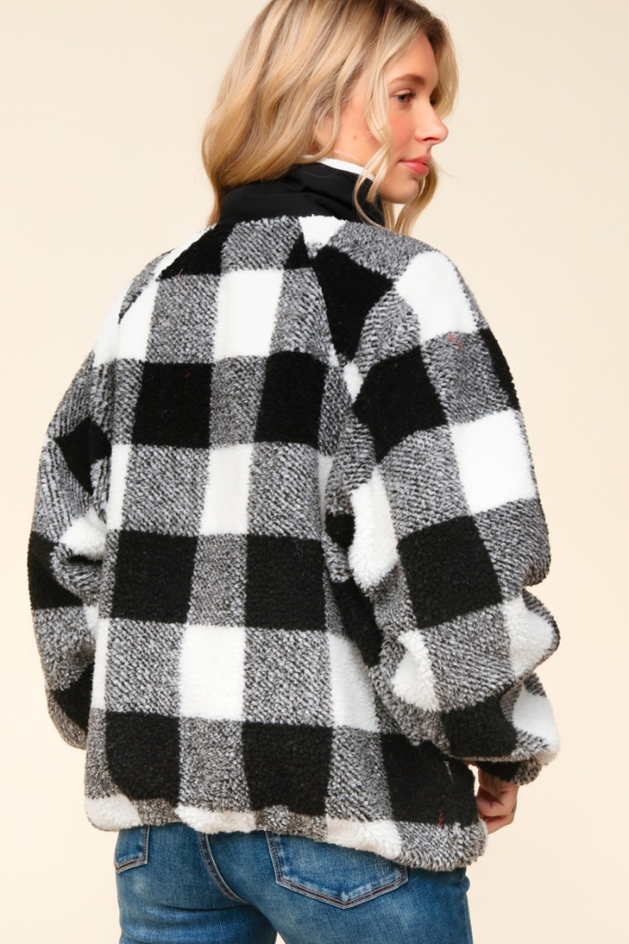 A woman from the back, with blonde hair, wearing a black and white buffalo plaid sherpa jacket with a black collar and zipper, and blue jeans