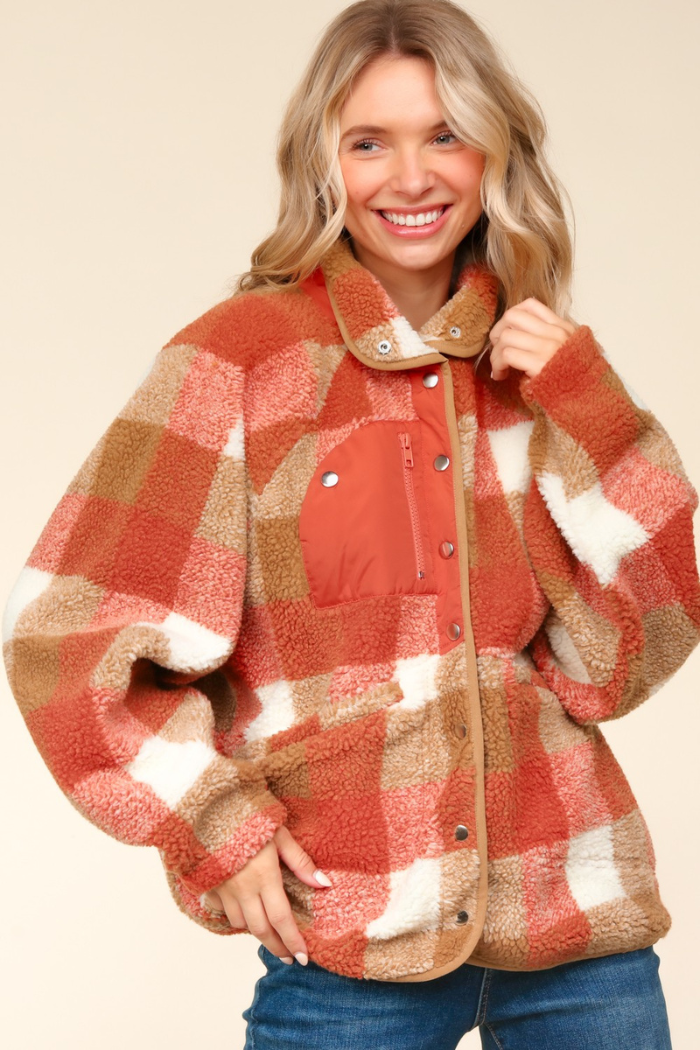 A smiling woman with blonde hair wears an oversized, rust and camel plaid sherpa jacket with white accents. The jacket features a high neck, a contrasting rust-colored chest zipper pocket, and a snap button-down front. She is wearing blue jeans and the background is a solid, light-colored wall. 