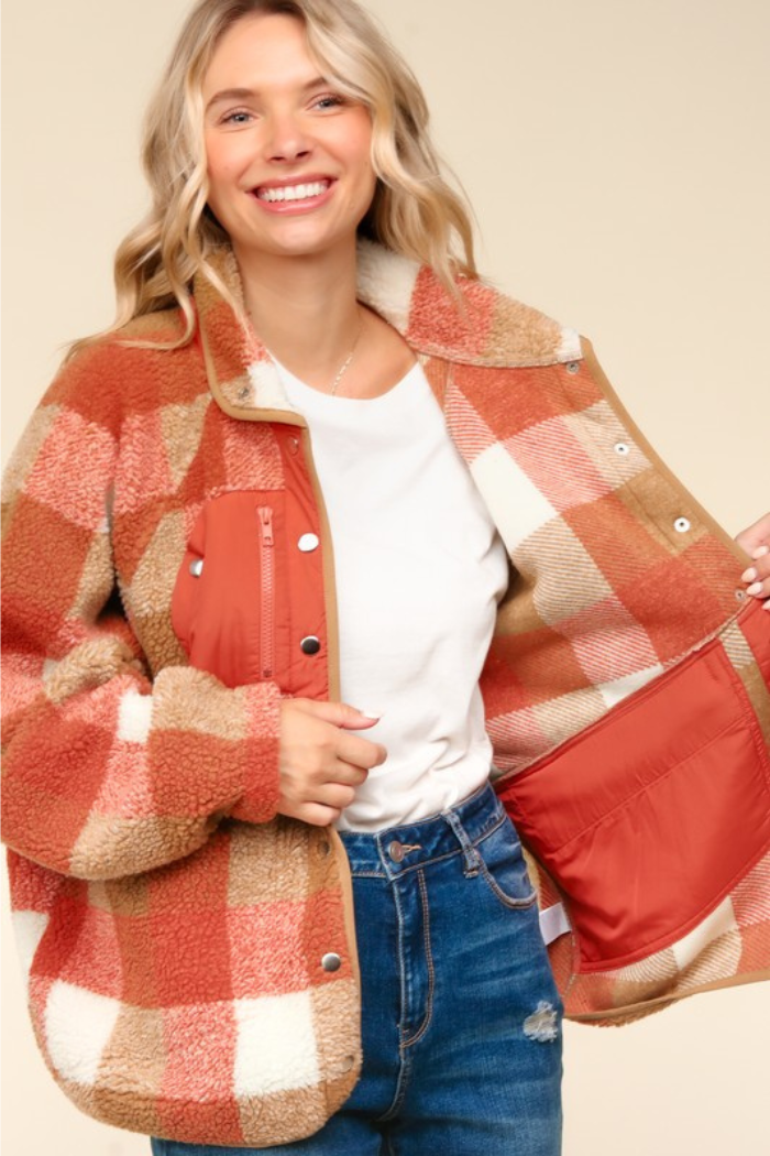 A woman with long blonde hair smiles while wearing an open, oversized, rust-colored and beige plaid sherpa jacket over a white t-shirt and blue jeans. The jacket has a zippered pocket on the left chest and snap buttons down the front. The inner lining of the jacket is a solid rust color. 