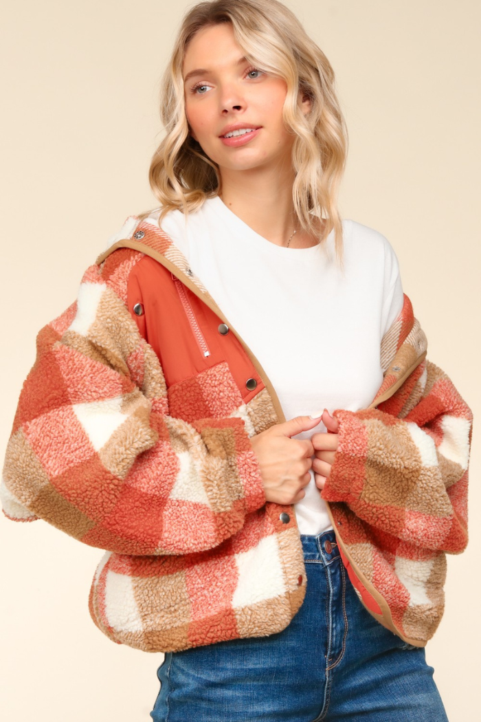 A young woman with blonde hair wearing a rust, camel, and white plaid sherpa jacket over a white t-shirt and blue jeans. She is looking slightly to her left and smiling softly, with her hands clasped in front of her. The background is a plain, light orange color.