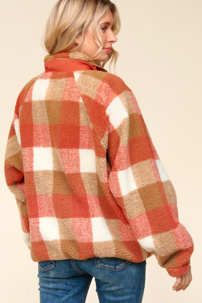 A woman from the chest up, seen from the back, wearing a rust, camel, and white plaid sherpa jacket with a high collar, over blue jeans. She has blonde hair and is looking over her right shoulder.
