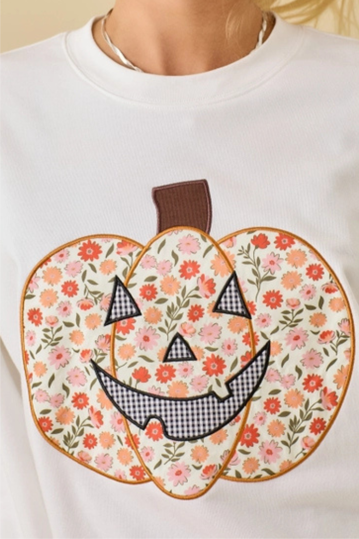 A close-up of a white long-sleeved shirt featuring a large, colorful floral and gingham patchwork pumpkin design. The pumpkin has a brown stem and a smiling jack-o'-lantern face with black and white gingham eyes and mouth. The body of the pumpkin is patterned with small pink and orange flowers on a cream background. 