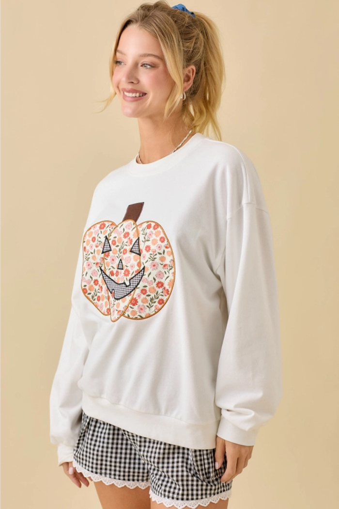 A young woman with blonde hair tied back in a ponytail smile while wearing a white long-sleeved sweatshirt featuring a large, colorful floral pumpkin design on the front. She is also wearing black and white checkered shorts with a white lace trim. The background is a plain light beige color.