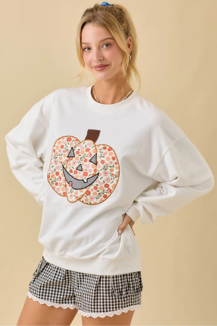 A young woman with blonde hair wearing a white crewneck sweatshirt featuring a large, colorful floral pumpkin graphic on the front. The pumpkin design incorporates various floral patterns and a black and white checkered pattern for the mouth, giving it a unique, patchwork-like appearance. The woman is also wearing black and white checkered shorts with a white lace trim. She is standing with her hands on her hips, looking directly at the viewer with a slight smile, against a plain, light-colored background.