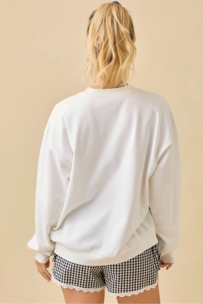 A person with blonde hair, pulled back in a ponytail, stands facing away from the camera, wearing an oversized white crewneck sweatshirt and black and white checkered shorts with a white lace trim.