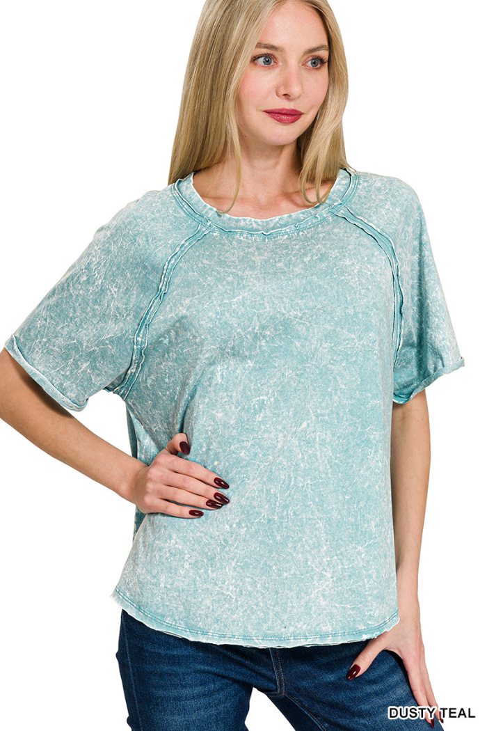 A woman wearing a dusty teal acid-washed short-sleeved top with raglan sleeves and a round neckline, standing with one hand on her hip against a white background.
