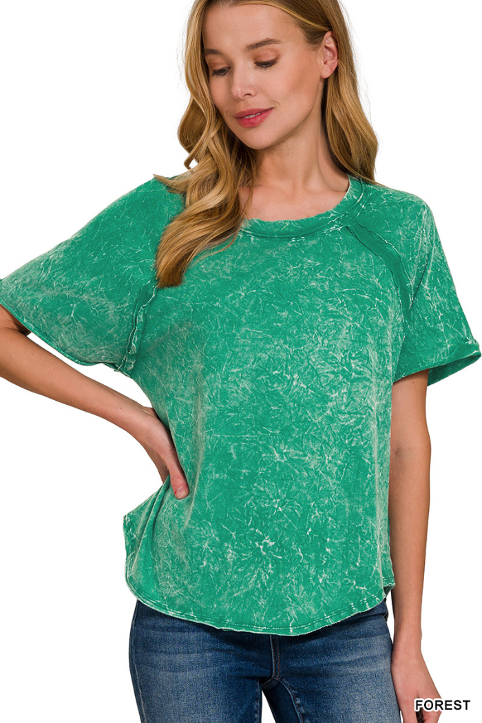 A woman wearing a green mineral wash short-sleeve top and jeans, with her left hand on her hip. The top has a textured, faded appearance. The background is plain white. The text "FOREST" is visible in the bottom right corner.