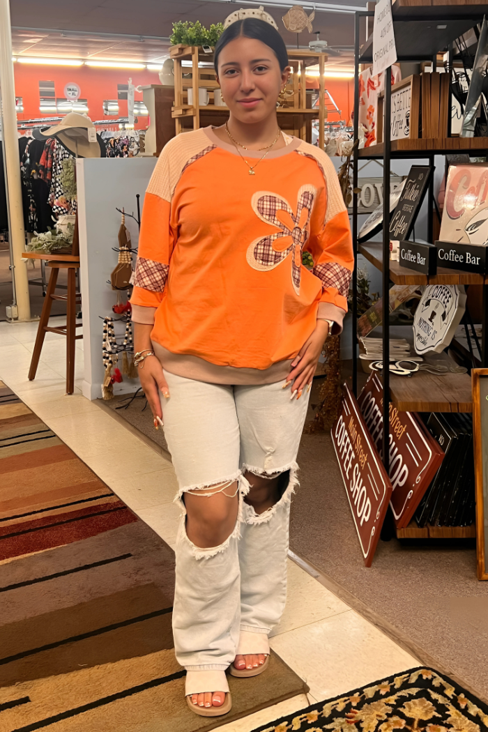 A young woman is wearing an orange and beige long-sleeved shirt with a large plaid flower design on the front, light-wash ripped jeans, and light-colored sandals. 